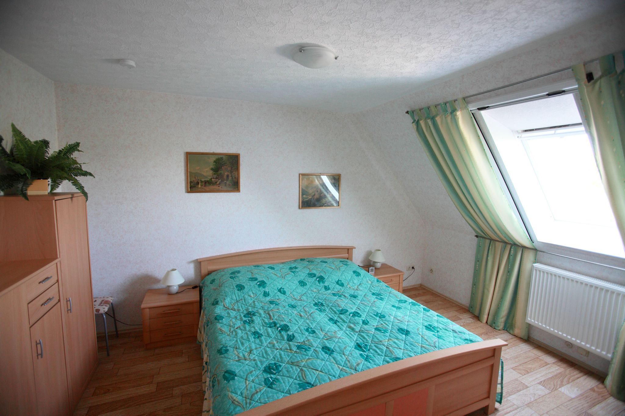 Een slaapkamer met een houten bed, een nachtkastje, een dressoir, een radiator en een raam met gordijnen.