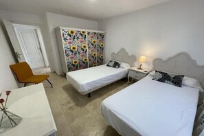 A bedroom with two single beds, a wardrobe with floral panels, a chair, a bedside table with a lamp, and a desk with a vase.
