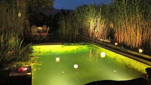 Zona de piscina exterior de noche con una mesa y sillas, rodeada de pastos altos y árboles.