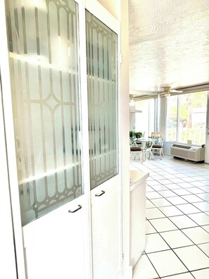 A room with a tiled floor, a white door with frosted glass panels, a ceiling fan, and a view of an outdoor patio area with chairs and a table.