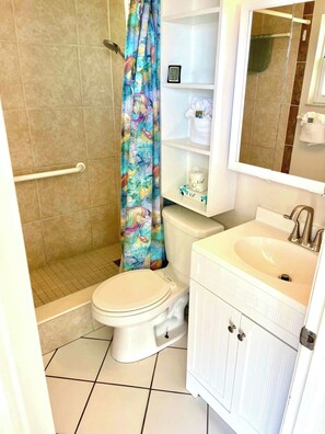 Ein Badezimmer mit Toilette, einer Dusche mit bunter Duschvorhang, einem Waschbecken mit Spiegel und Ablage für Toilettenartikel.