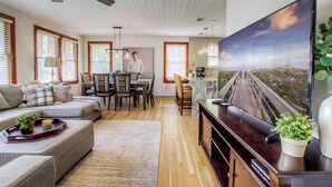 Una sala de estar moderna con sofá, comedor y una gran pantalla de televisión que muestra un paisaje.