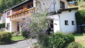 A two-story building with a balcony adorned with flowers, a garden with purple flowers, and a mountain backdrop.