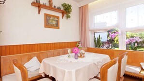 Uma área de refeições com uma mesa redonda coberta por uma toalha branca, rodeada por cadeiras de madeira com almofadas brancas. Há um banco de madeira ao longo da parede, uma estante com objetos decorativos e uma janela com vista para árvores e flores.