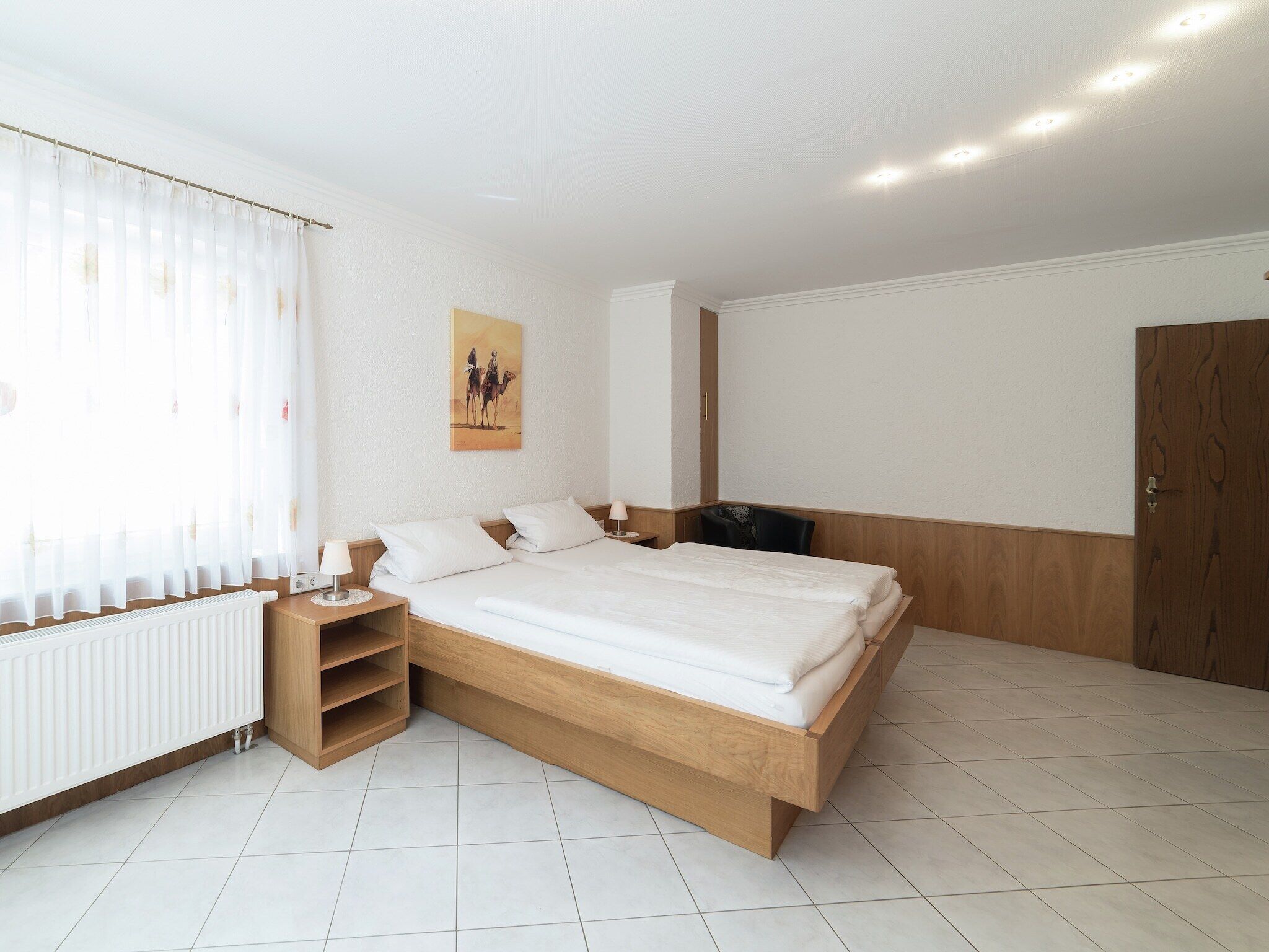 Un dormitorio con cama de madera, una mesita de noche, un radiador y una ventana con cortinas translúcidas.