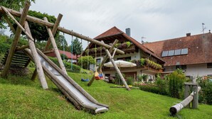 A playground with a slide and swings, surrounded by a well-maintained lawn and a house with a red-tiled roof. - Schwarzwald-Ferienwohnung auf dem Bauernhof (Schuttertal-Regelsbach)