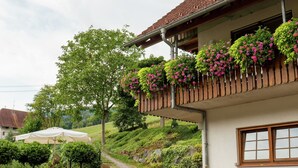 A building with a balcony adorned with hanging flower baskets, a paved pathway leading to a covered seating area, and a view of a hillside. - Schwarzwald-Ferienwohnung auf dem Bauernhof (Schuttertal-Regelsbach)