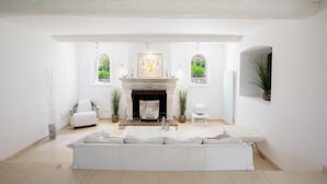 Amplia sala de estar con chimenea, sofás blancos y vistas a follaje a través de ventanales abovedados.