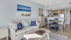 A modern living room with a beige sofa, a coffee table, and a dining area with wooden chairs and a table. A ceiling fan is visible, and there is a kitchen area with cabinets and a microwave.