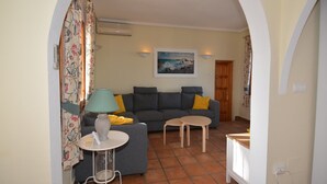 A living room with a grey sofa, a small round table, and a lamp. There is a painting on the wall and a door in the background.
