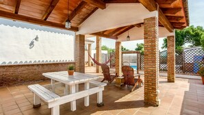 Une terrasse couverte avec une table et des chaises en bois, un hamac et une vue sur une piscine.