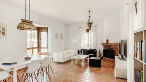 Un salon spacieux avec un canapé blanc, un canapé noir et une table basse. On y trouve une cheminée avec un manteau en briques et un téléviseur posé sur un meuble blanc. La pièce est dotée d’un sol carrelé et comprend un coin repas avec une table et des chaises blanches.