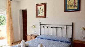 Una cama bien hecha con una colcha azul estampada y dos toallas enrolladas. Junto a la cama, una mesita de noche de madera con una lámpara.