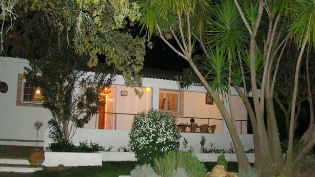 Un edificio blanco con un balcón, rodeado de un jardín con plantas variadas y una palmera.