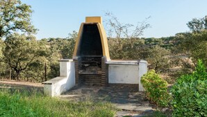 Un arco de piedra que da acceso a una construcción de ladrillo con chimenea, rodeada de follaje y árboles.