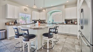 Una cocina moderna con armarios blancos, una isla grande con taburetes de bar, electrodomésticos de acero inoxidable e iluminación colgante.
