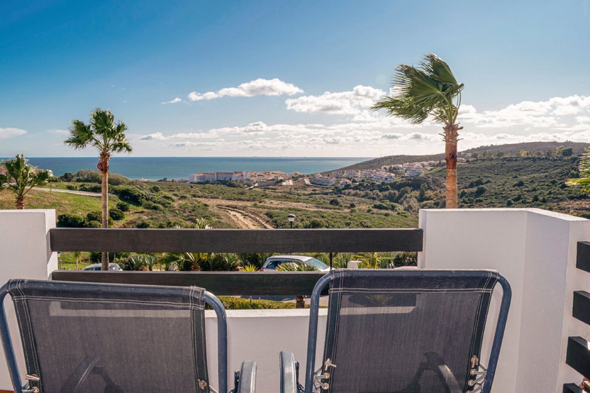 A balcony with two chairs facing a coastal view, palm trees, and a distant town.