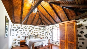 Un dormitorio con pared de piedra, vigas de madera en el techo, una cama con un banco y un armario de madera.