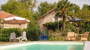 A swimming pool with lounge chairs and umbrellas, surrounded by greenery and a stone building.
