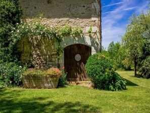 En rustik steng bygning med en trædør, omgivet af frodigt grønt og en velplejet græsplæne.