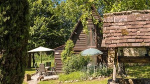 A rustic wooden building with a stone wall, a stone pathway, and outdoor seating with umbrellas. - Rustic Chic Country Cottage (Thenon)