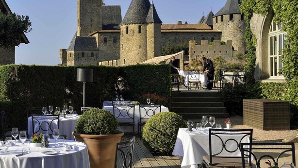 Un área de comedor al aire libre con mesas dispuestas para una comida, rodeada de vegetación y un castillo al fondo.
