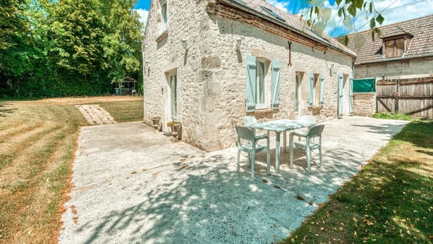 Ein Steinhaus mit Ziegeldach, einem kleinen Garten und einer Terrasse mit Tisch und Stühlen.