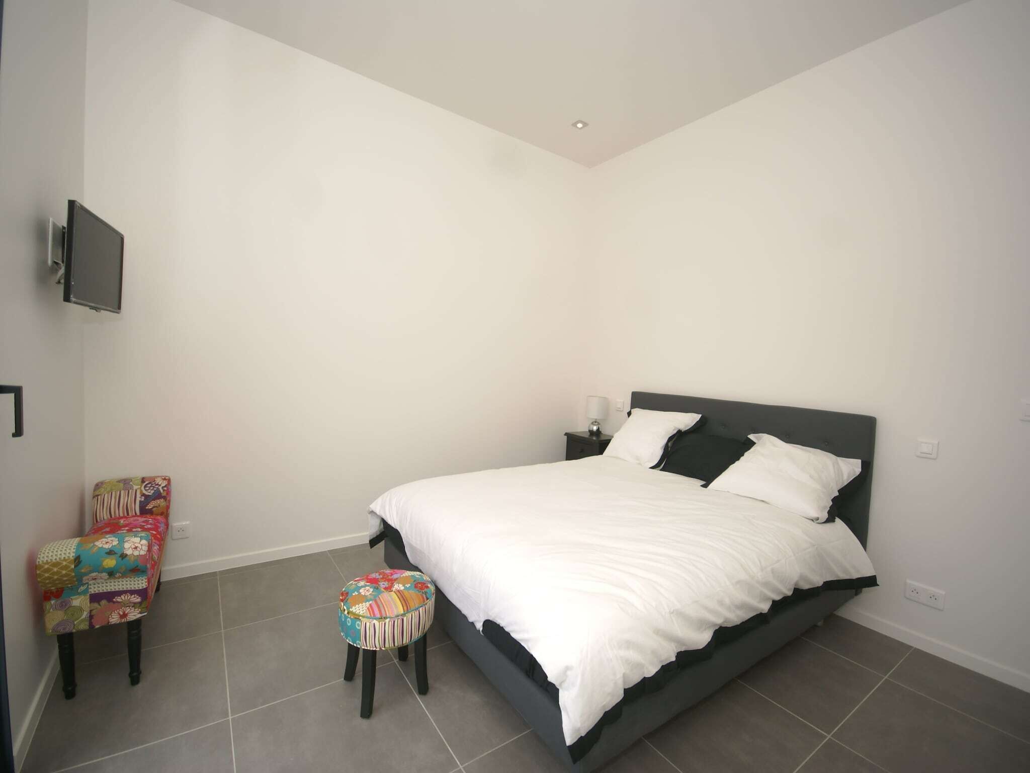 A bedroom with a bed, a small stool, a chair, a bedside table, and a TV mounted on the wall.