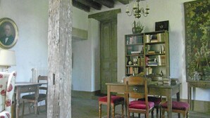 Ein rustikaler Raum mit sichtbaren Holzbalken, einer Chandelieber, einem Bücherregal, einem Esstisch mit Stühlen und einem gerahmten Porträt an der Wand.