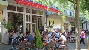 Ein Straßencafé mit Korbstühlen und Tischen, einem großen Topfpflanzen und einem roten Sonnenschirm mit Beschilderung.
