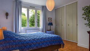 Une chambre à coucher avec un grand lit, une fenêtre avec des rideaux, une lampe de chevet, une armoire et un luminaire suspendu.