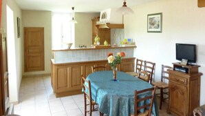 A dining area with a table set for four, wooden chairs, a blue tablecloth, a vase with flowers, a kitchen area with wooden cabinets, a hanging light fixture, and a television on a wooden cabinet.