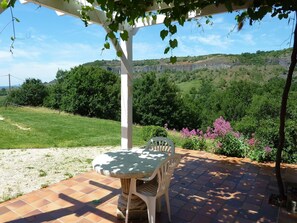 Um pátio externo com mesa e cadeiras, uma pérgula e vista para uma paisagem verde com colinas.