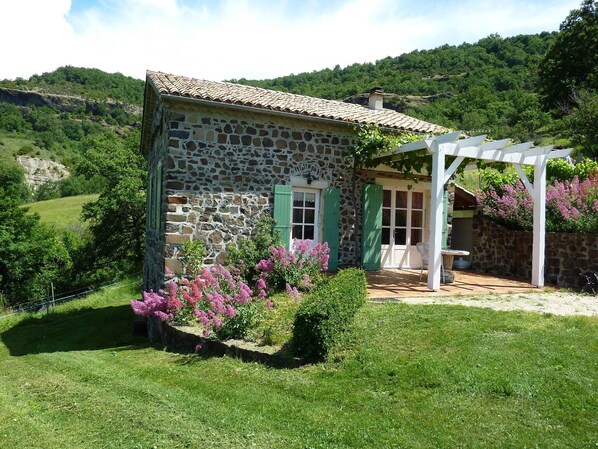 Uma casa de pedra com telhado de telhas, pérgola de madeira e um jardim com flores cor-de-rosa.