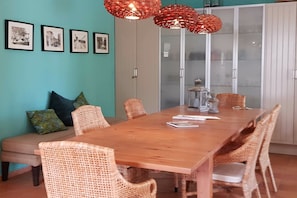 Un coin repas avec une table en bois, des chaises en osier et un banc. On y trouve des tableaux encadrés au mur, un meuble avec des portes en verre et un luminaire rouge suspendu.