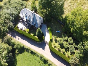 An aerial view of a large, two-story house with a stone facade, surrounded by well-maintained gardens and a curved driveway.