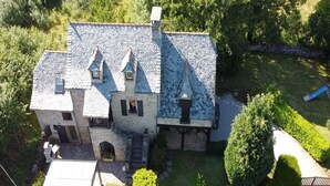 Une maison en pierre avec un toit à forte pente, plusieurs cheminées et une petite dépendance attenante dotée d’un balcon.