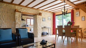 Uma sala de estar acolhedora com parede de pedra, vigas de madeira, um sofá, uma mesa de centro e um espaço de jantar com cadeiras de vime.