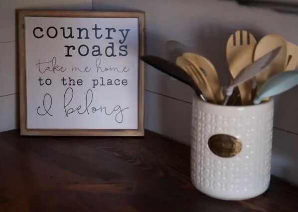 Uma placa emoldurada com o texto 'country roads take me home to the place I belong' e uma caneca de cerâmica branca com utensílios de madeira.