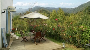 A wooden deck with a table and chairs, an umbrella, and a view of a lush garden and mountains. - Riverside Home in Folelli (Folelli)