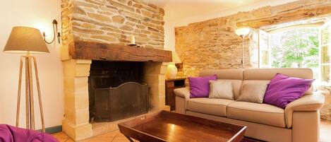 Una sala de estar con chimenea de piedra, un sofá color beige con cojines morados, una mesa de centro de madera y una alfombra morada.