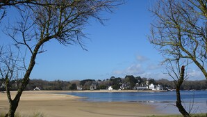 A coastal landscape with a sandy beach, grassy areas, and a row of houses along the water's edge. - Cottage (Guidel)