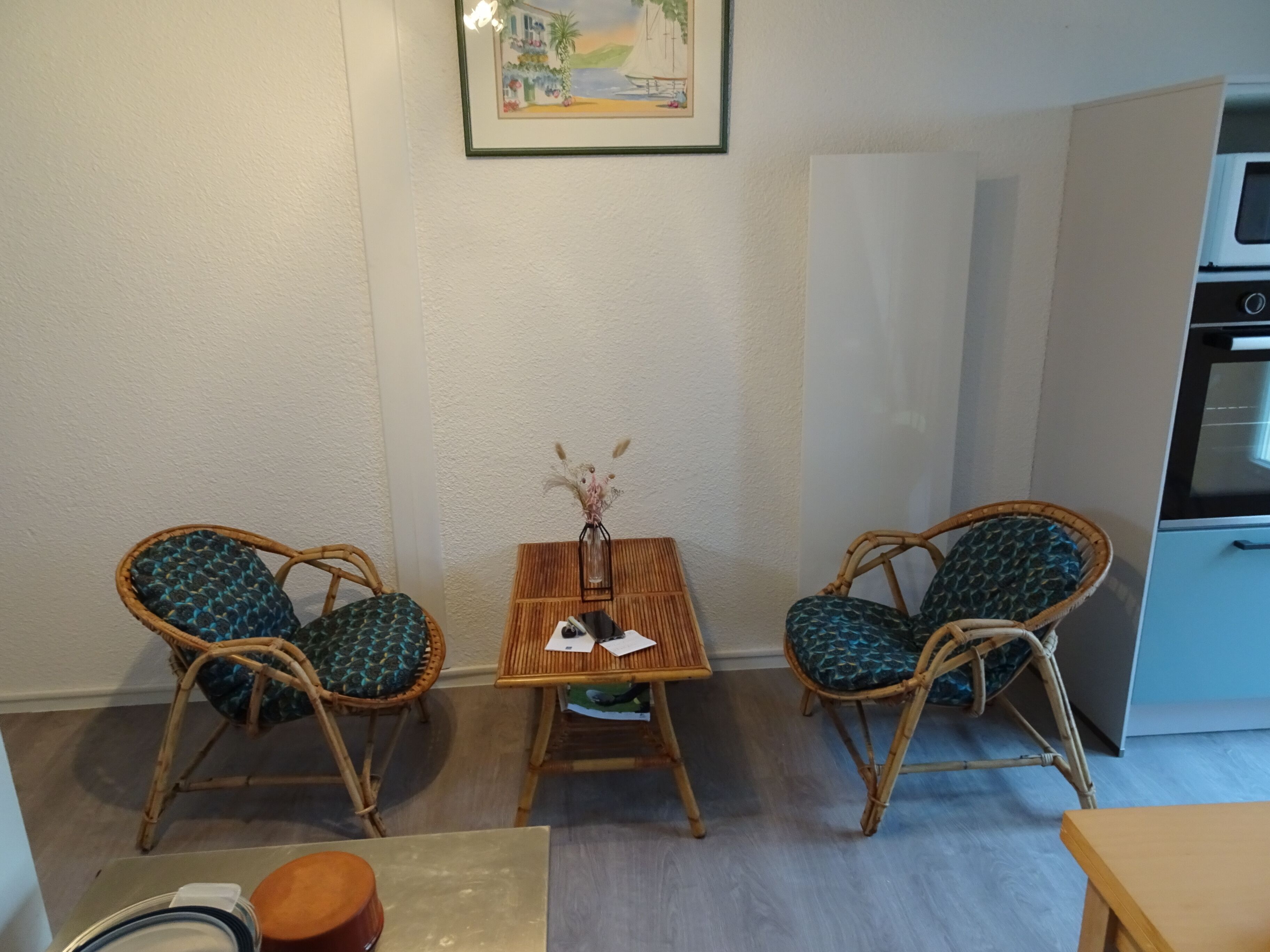 A small, cozy room with two wicker chairs, a wooden table, and a framed picture on the wall.