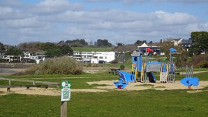 A playground with various play structures, a sign indicating a tobacco-free area, and a view of houses and a beach in the background. - Cottage (Guidel)