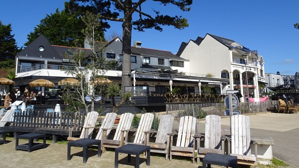 A seaside restaurant with outdoor seating, a large tree, and a building with a balcony. - Cottage (Guidel)