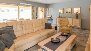 A modern living room with a beige sofa, a wooden coffee table, and a leather armchair. There is a dining area visible through the window. - Pleasant holiday home with private pool (St Nexans)
