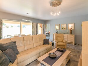 A modern living room with a sofa, armchair, and a dining table. There are large windows, a ceiling light fixture, and a small cabinet. - Pleasant holiday home with private pool (St Nexans)