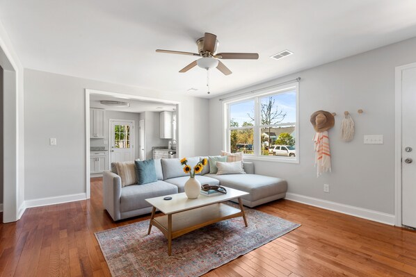 A spacious living room with hardwood flooring, a ceiling fan, a grey sofa, a coffee table with a vase of sunflowers, and a view of the outside through large windows.