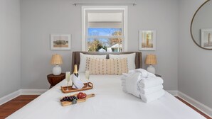 A neatly arranged bedroom with a large bed, two bedside tables with lamps, a window with a view, and framed pictures on the walls.
