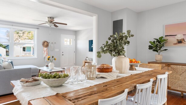 A spacious living room with a wooden dining table, white chairs, a grey sofa, and a ceiling fan.
